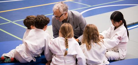Ein Trainer spricht mit einer Gruppe von fünf Kindern in Judoanzügen auf einer Sportmatte.